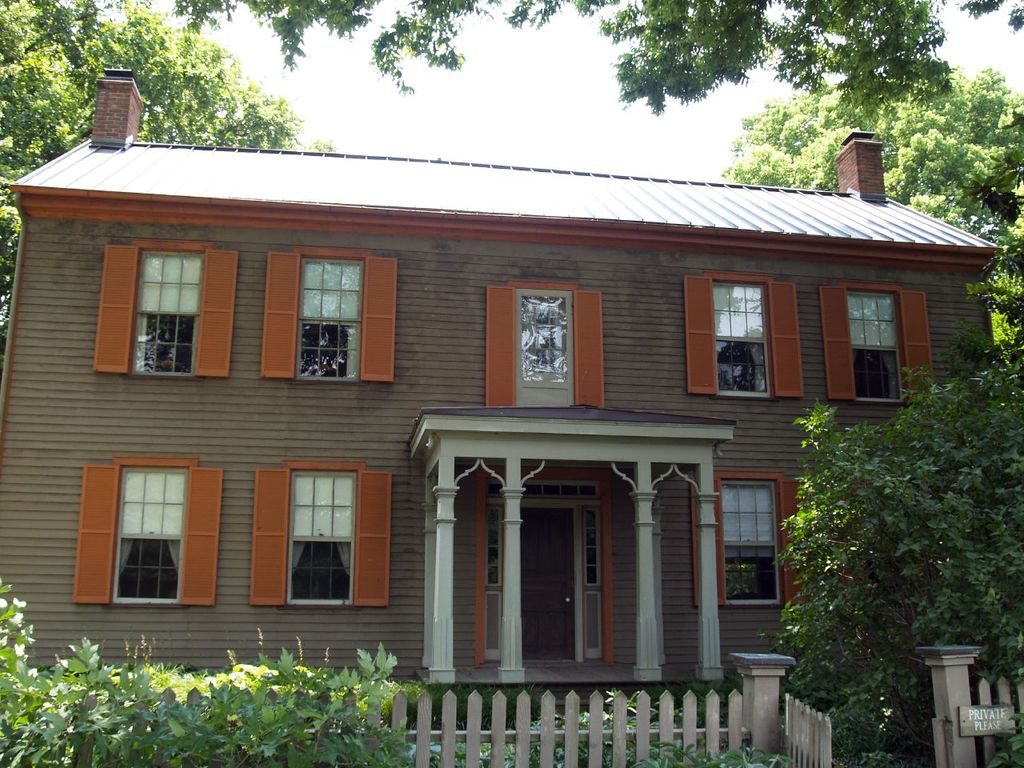 Two storey wooden house with orange shutters, a columned porch and a white picket fence